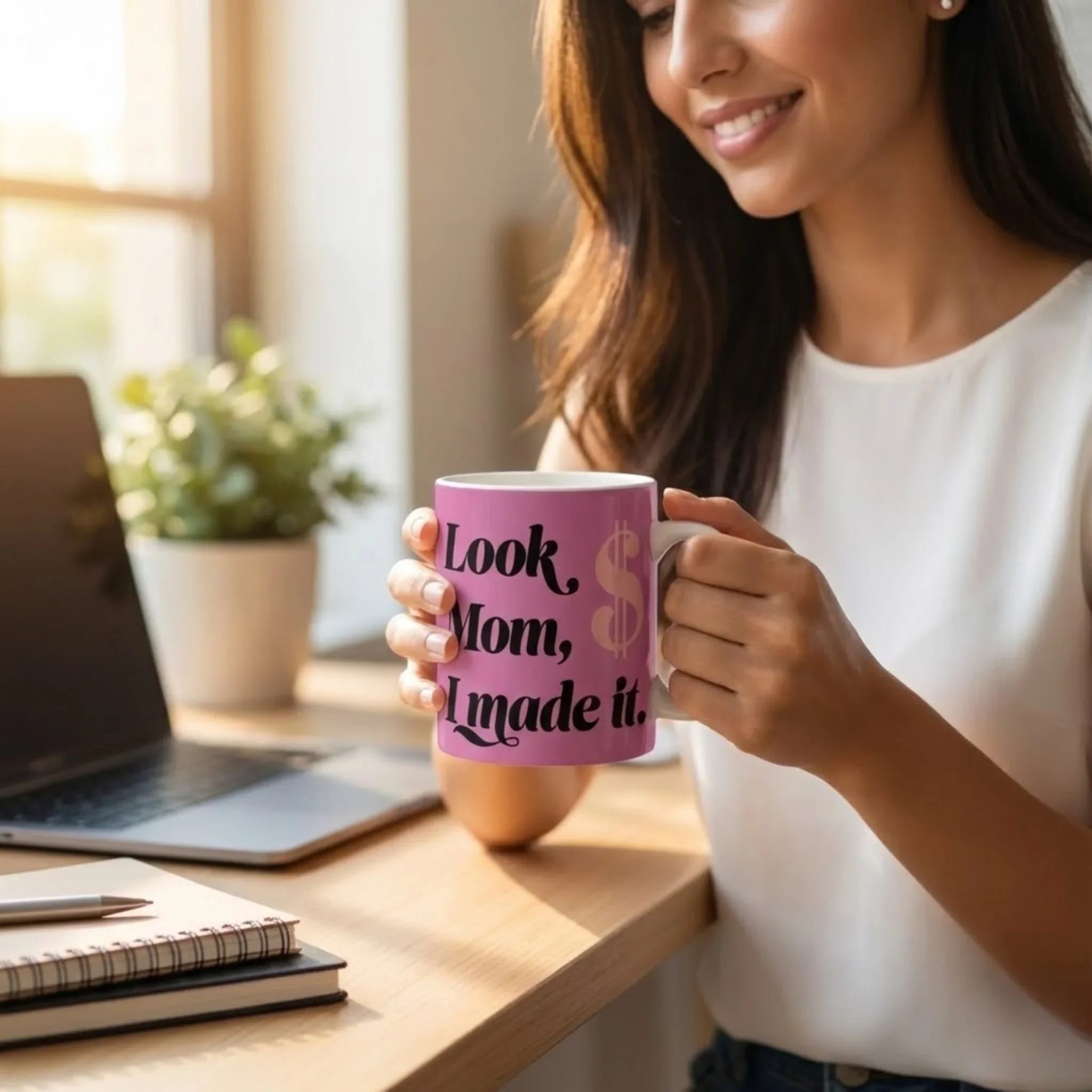 Woman holding a pink mug with text, sitting at a desk with a laptop and notebook.