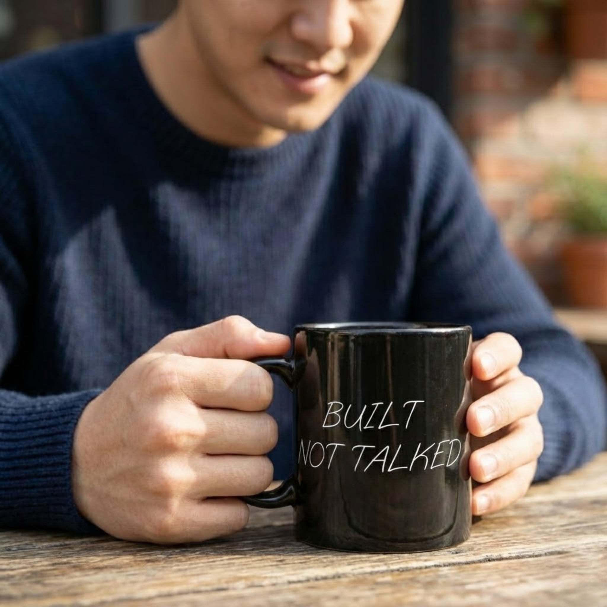 Person holding a black mug with 'BUILT NOT TALKED' text on a wooden surface