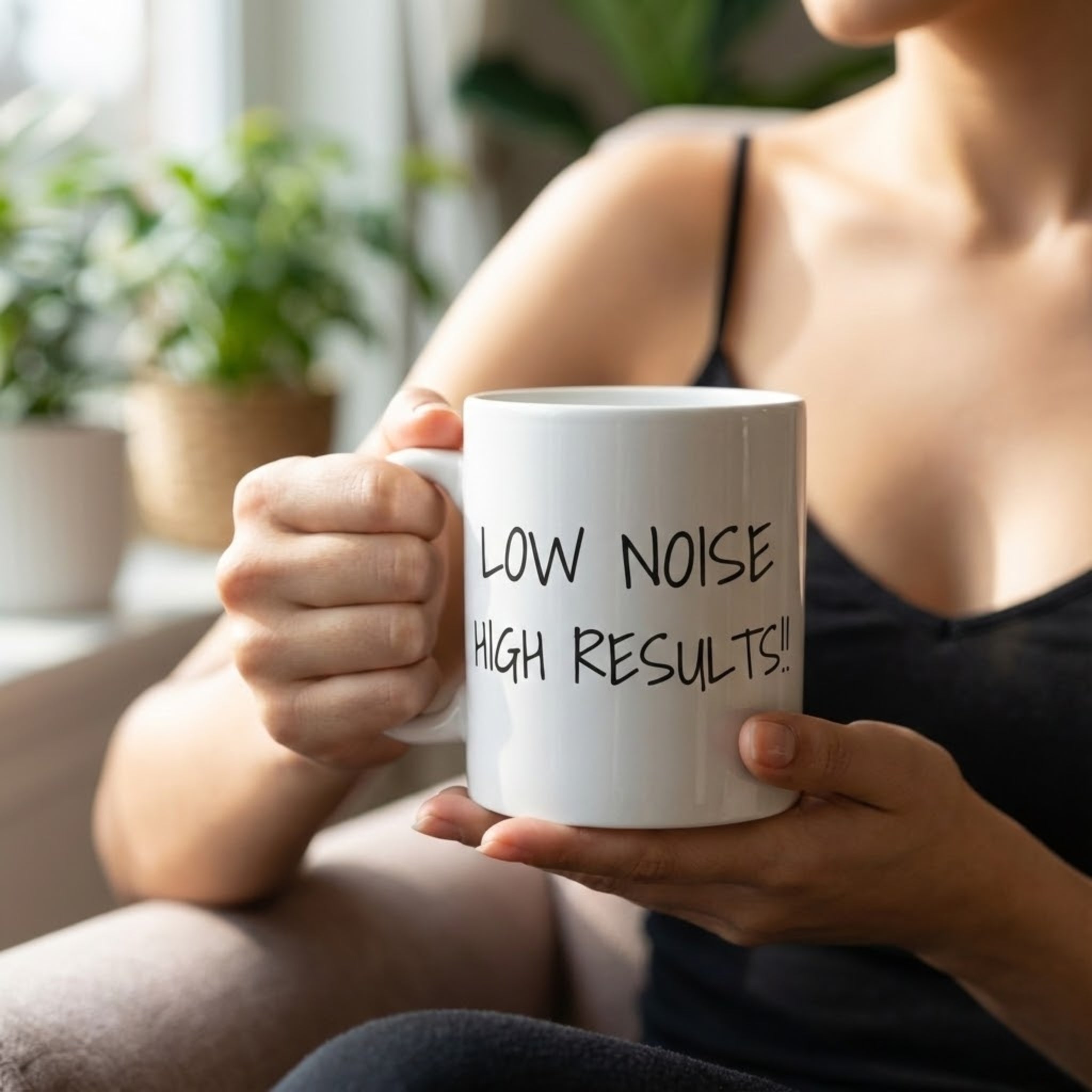 Person holding a white mug with 'LOW NOISE HIGH RESULTS!' text in a home setting.